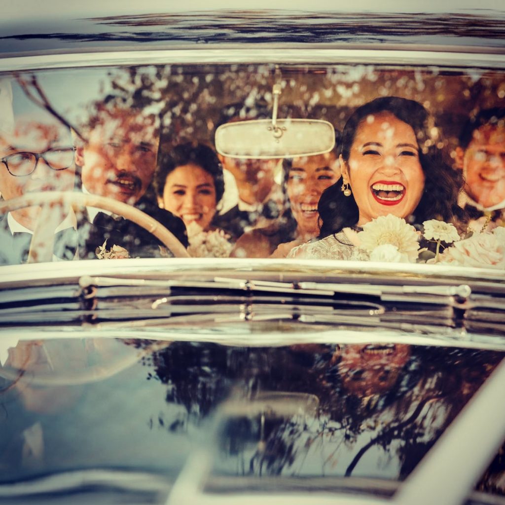 A laughing bridal party riding in a vintage Humber, seen through the windscreen